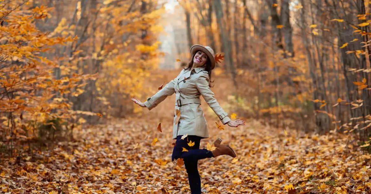 Woman joyfully jumping in a forest covered with autumn leaves, wearing a beige trench coat, hat, and boots during a cozy fall weekend getaway.