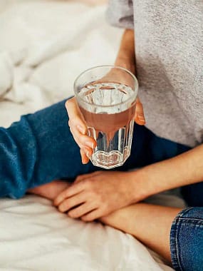 drinking water-8-2 woman sitting on bed holding a glass of water as one of many tricks to help hydrate better