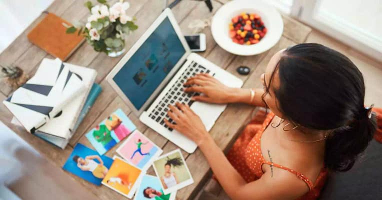 blog post ideas woman sitting at a wooden table working on her laptop researching ways to create residual income surrounded by colorful candy in a white bowl, Polaroid photos, notebooks, and flower in clear vase.