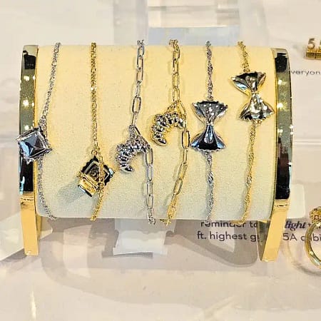 a selection of gold and silver bracelets displayed on a stand inside a jewelry case representing different items that include flaky croissants and bowtie pasta.