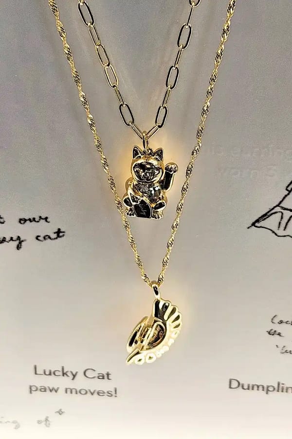 two gold necklaces layered vertically. necklace on top is a cute cat waving and the one on the bottom is a dumpling.