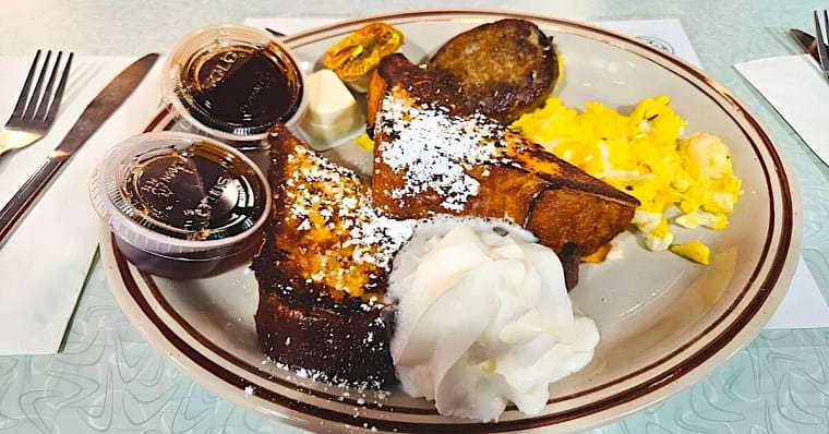 1-4 comfort breakfast food featuring French toast sprinkled with powered sugar, scrambled eggs, whipped cream, sides of maple pancake syrup ins plastic container and sausage patty on a plate.