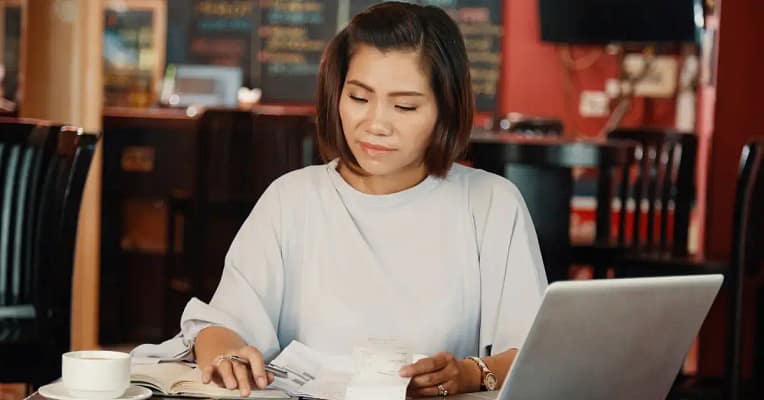 saving money budgeting woman sitting in a shop wearing a quarter sleeve shirt and a gold watch sitting at a table with laptop, coffee, book, papers, and ink pen budgeting her finances.