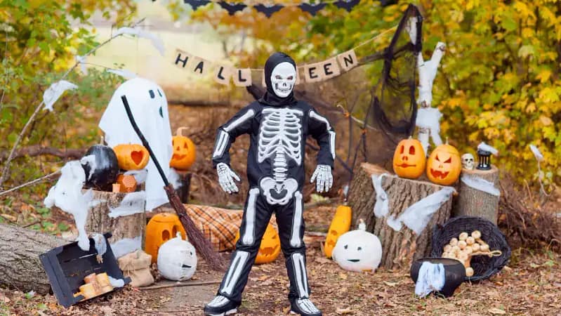 a little kid standing outside dressed in a simple skeleton bodysuit with halloween decorations in the background behind them.