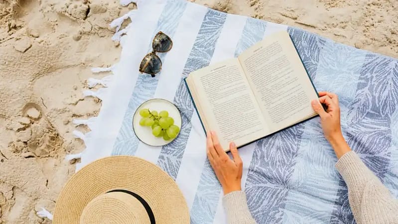 A person sprawled on a beach towel, reading a book while soaking up the sun with sunglasses and grapes sitting in front of them.