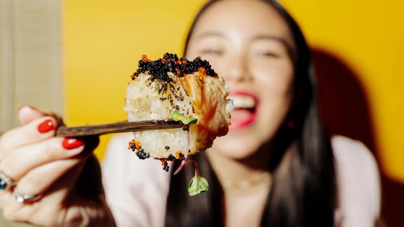 young asian woman holding a sushi roll with brown wooden chopsticks wearing red nail polish
