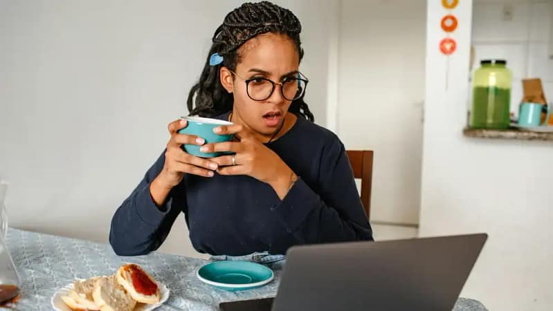 black woman with braids and glasses sitting at the table looking a computer screen in shock while holding a light blue ceramic cup with both hands