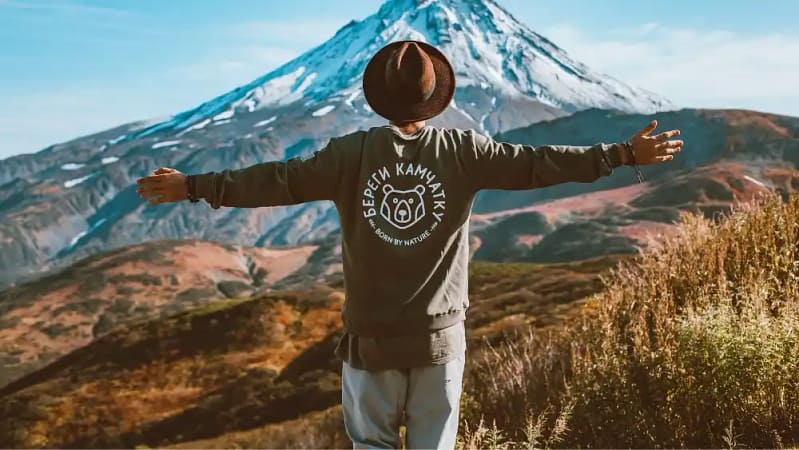 white man standing straight with both arms stretched out side to side looking at a mountain covered in snow or ice