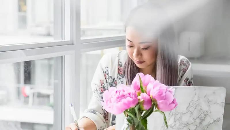 a picture of a young asian woman with lavendaire hair sitting at a desk using an ink pen