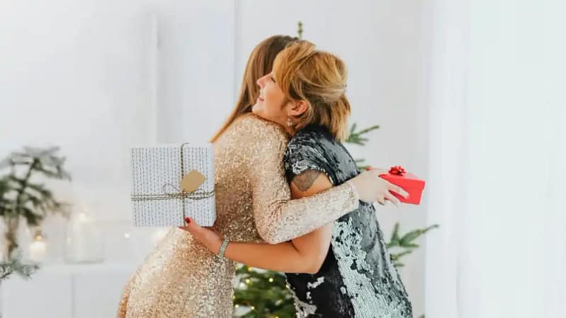 two women embracing in a warm hug both holding a gift and wearing sequin dresses