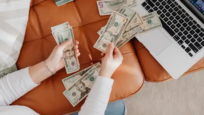 woman sitting on a light brown leather couch in front of a laptop organizing a pile of money