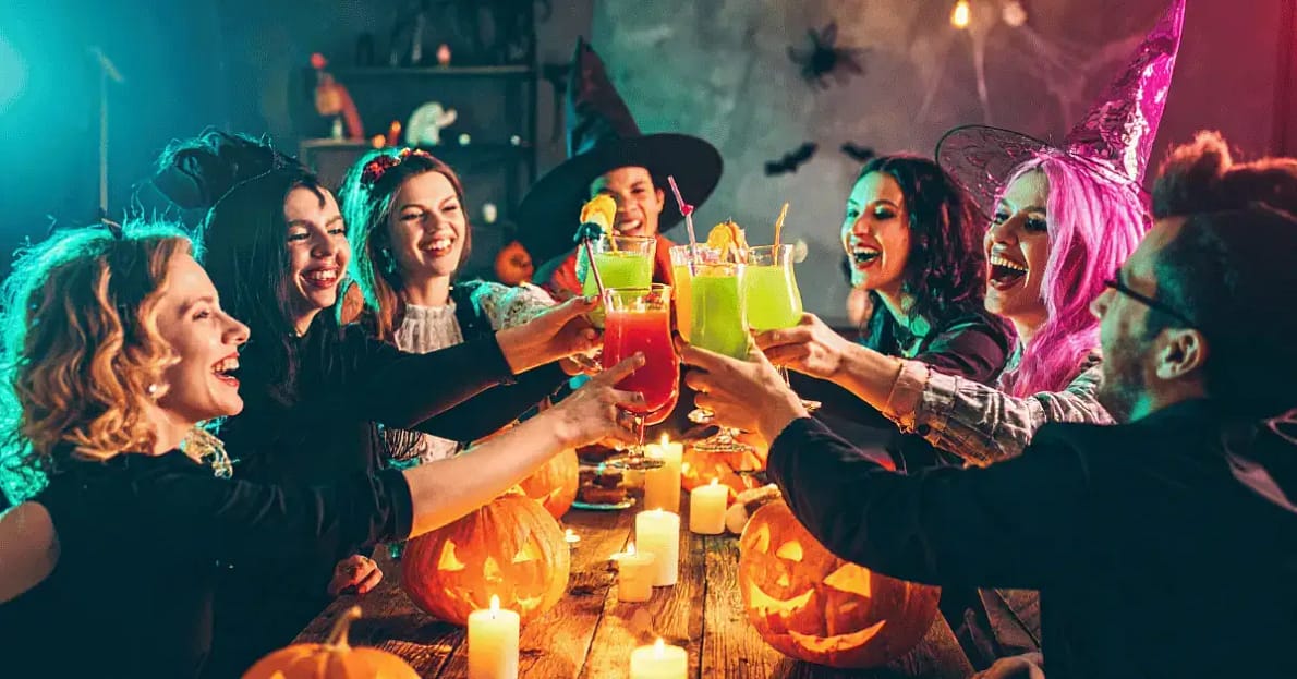 Group of friends in Halloween costumes celebrating with colorful drinks, jack-o’-lanterns, and candles at a festive Halloween party.