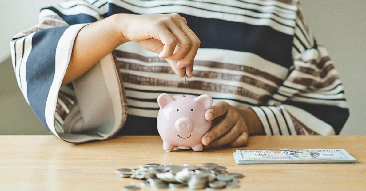 person sitting at a table surrounded by silver coins and 100 dollar bills putting coins inside of a small pink piggy bank