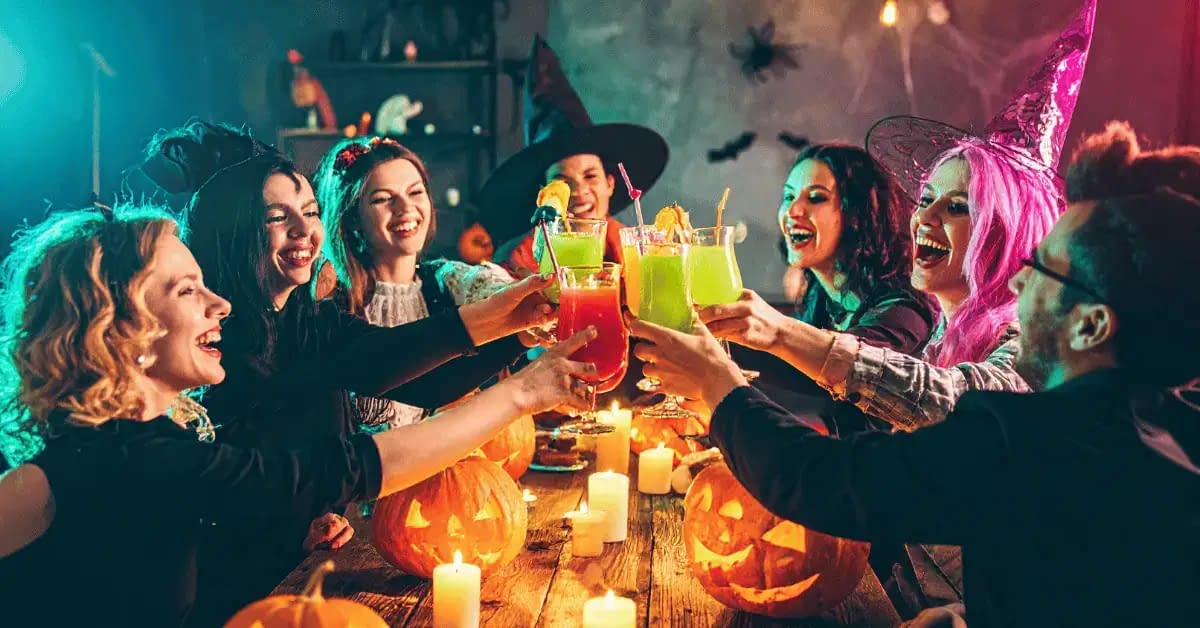 Group of friends in Halloween costumes celebrating with colorful drinks, jack-o’-lanterns, and candles at a festive Halloween party.