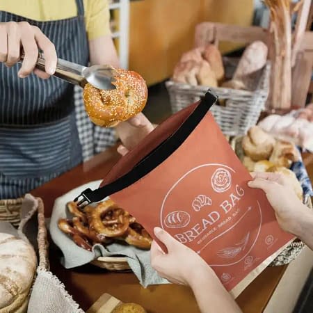 Person placing a sesame bagel into an eco-friendly reusable bread bag at a bakery counter.