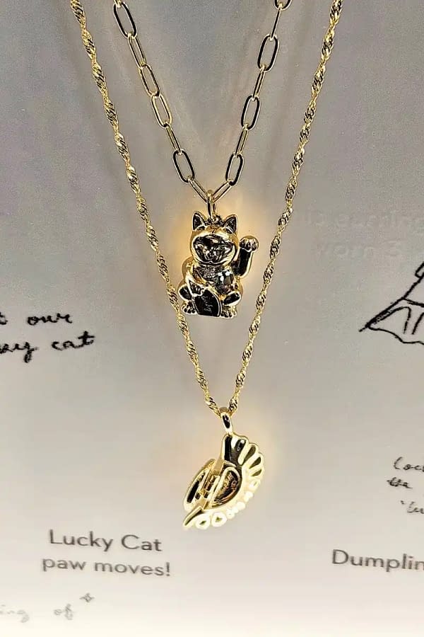 two gold necklaces layered vertically. necklace on top is a cute cat waving and the one on the bottom is a dumpling.