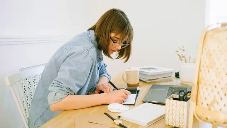 woman writing down blog post ideas in a journal notebook while sitting in a white chair at a long wooden desk