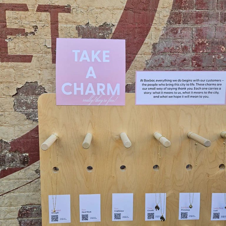 a wooden wall of bracelet charms and charm necklace saying "Take A Charm".
