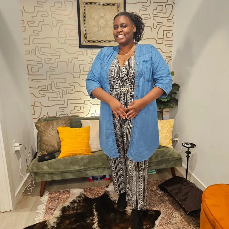 Philly blogger wearing a white and black jumpsuit with a printed pattern, a light washed denim jacket and black suede boots standing in a cozy setting