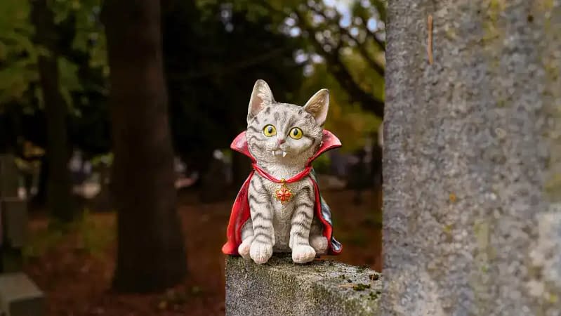 striped cat statue or ornament placed on a ledge outside wearing a red and black vampire cape, a red and gold necklace with fangs sticking out it's mouth.